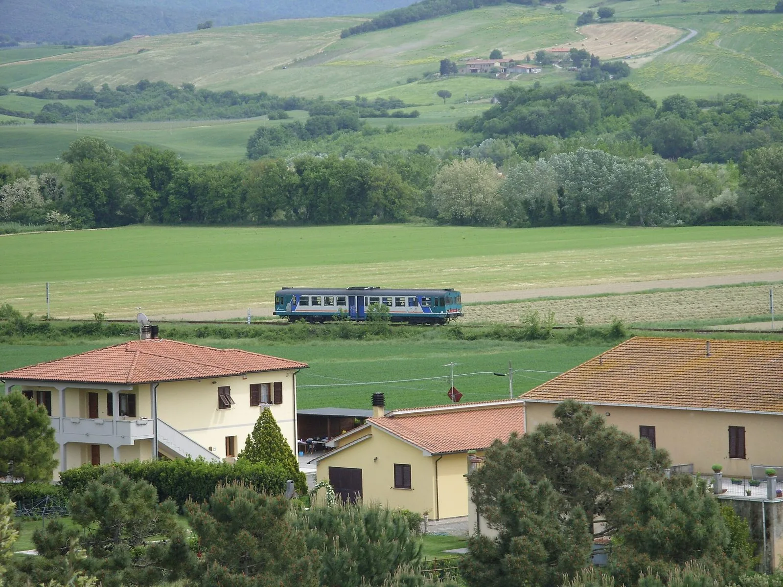 Automotrice regionale che attraversa i campi verdi della Val di Cecina lungo la ferrovia Cecina-Volterra, con colline e casali sullo sfondo